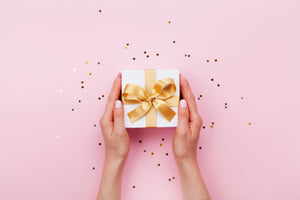Hands holding a white gift box with gold ribbon agains a light pink background.
